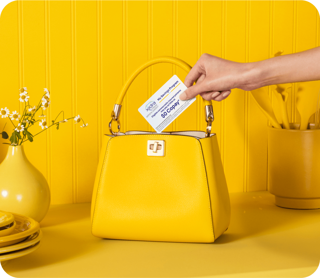 Hand holding a Xiidra Savings Program card above a yellow purse on a yellow counter