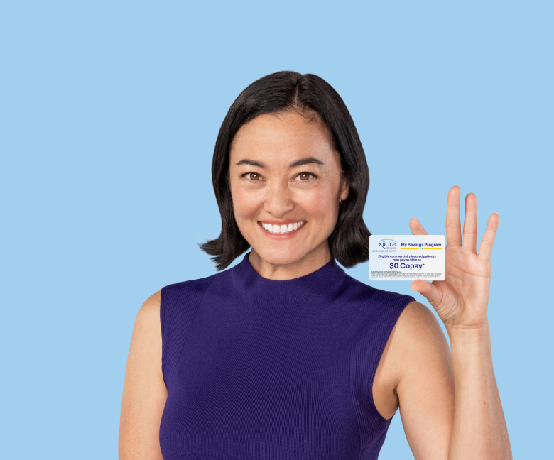 Woman with short hair in a purple top holds her Xiidra Savings Program card up